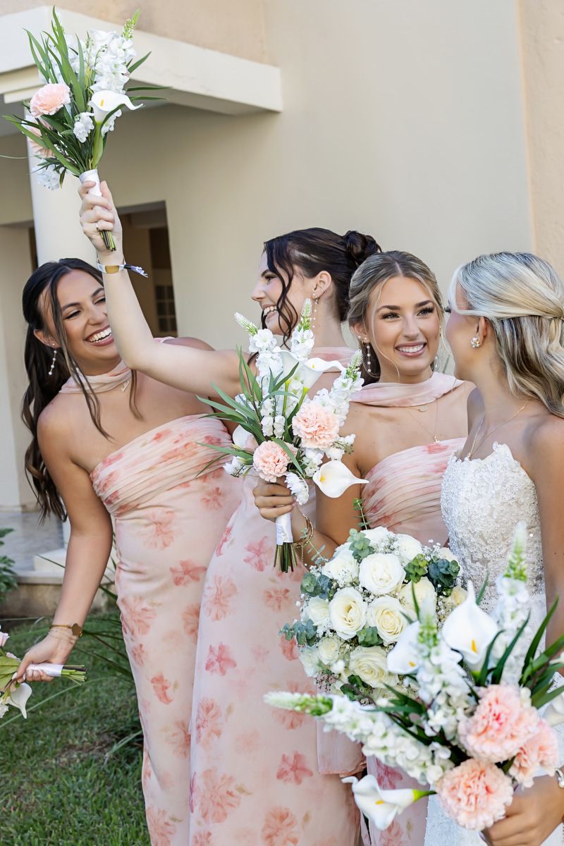 Bridesmaids in pink floral dresses holding bouquets of white and pink flowers, laughing and celebrating outdoors.
