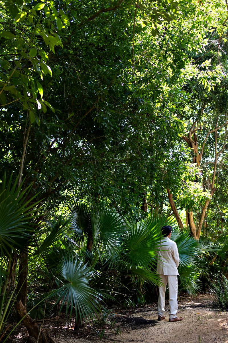 Person in white outfit standing on a dirt path surrounded by lush tropical greenery and palm trees in a forest.