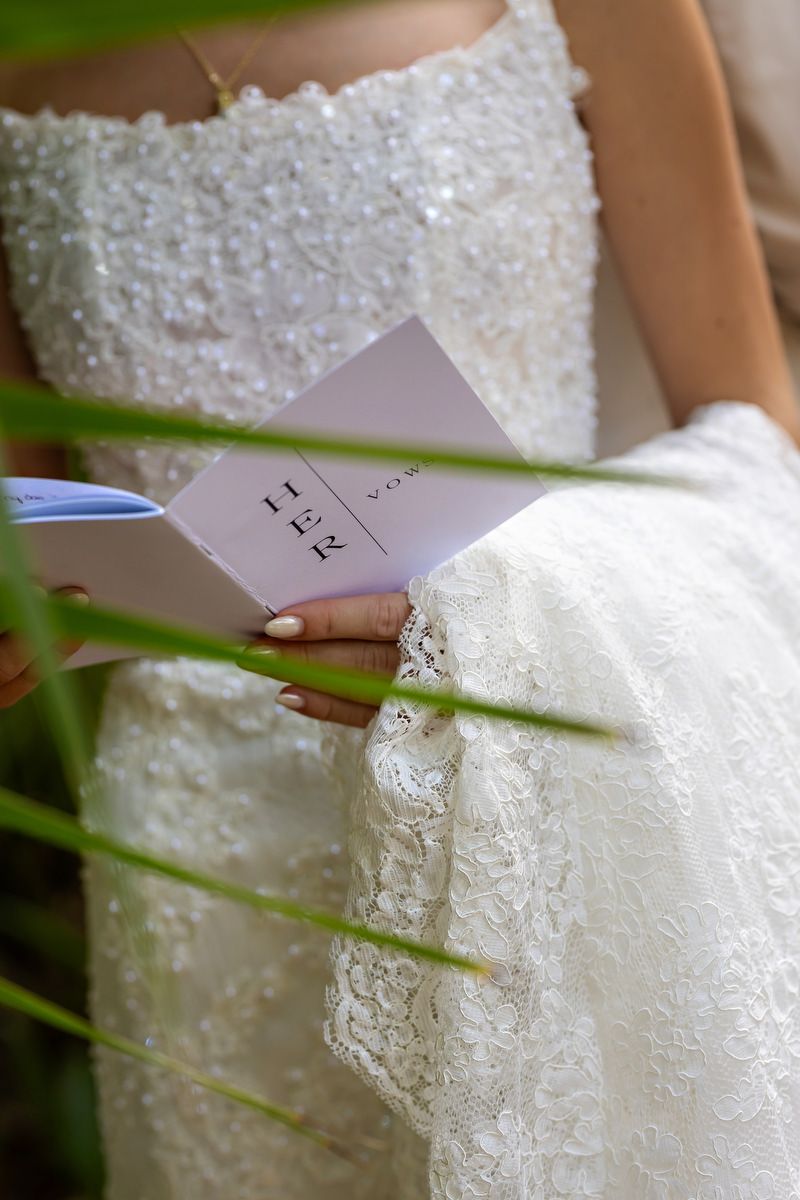 Bride in lace wedding dress holding vow booklet outdoors, elegant beaded bodice detail visible.