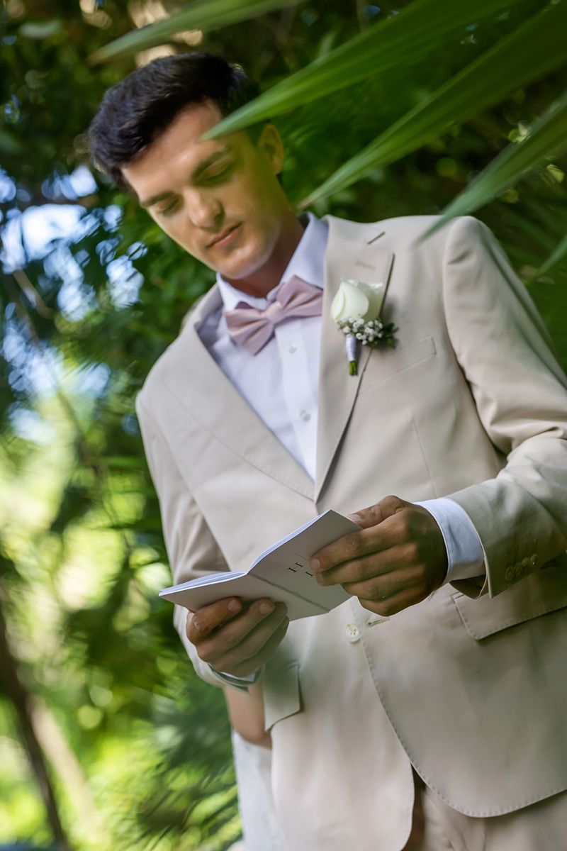 Groom in beige suit with pink bow tie and boutonniere reads wedding vows outdoors among tropical greenery.