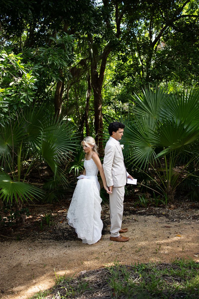 Bride in white dress and groom in tan suit stand back-to-back among lush tropical palm trees during first look.