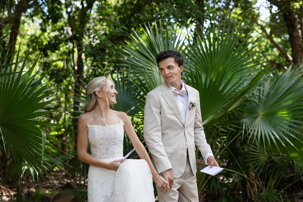 Smiling bride and groom hold hands among tropical palms, groom holding vows card during outdoor wedding ceremony.