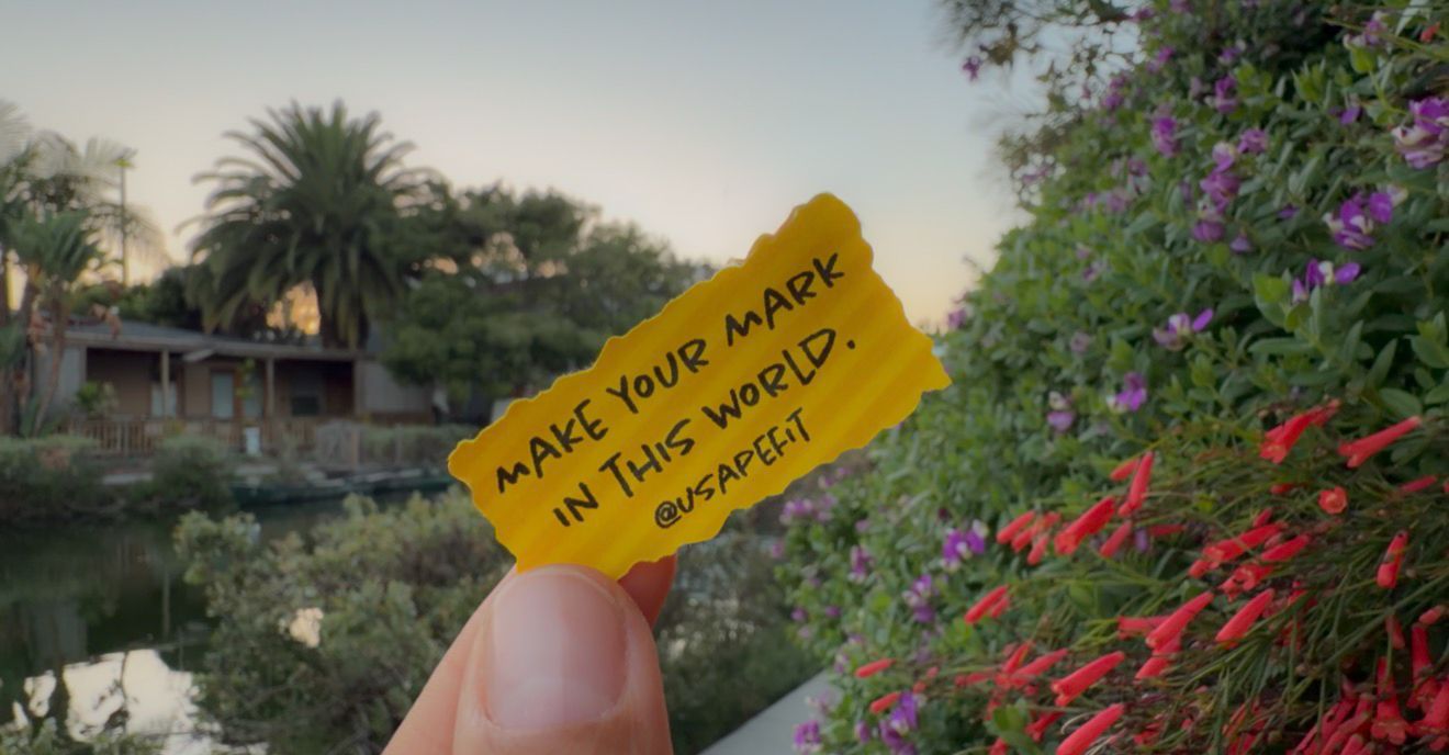 Hand holding a yellow paper note reading Make Your Mark In This World outdoors near water and purple flowers.