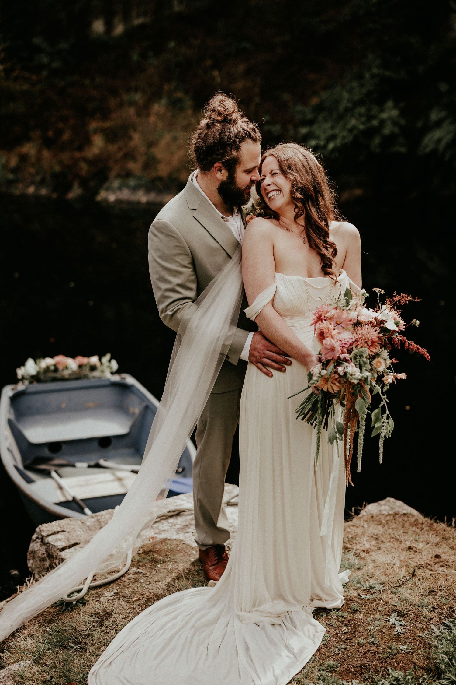 Bride and groom embrace lakeside, she holds a colorful bouquet in a white gown, he wears a tan suit, rowboat behind them.