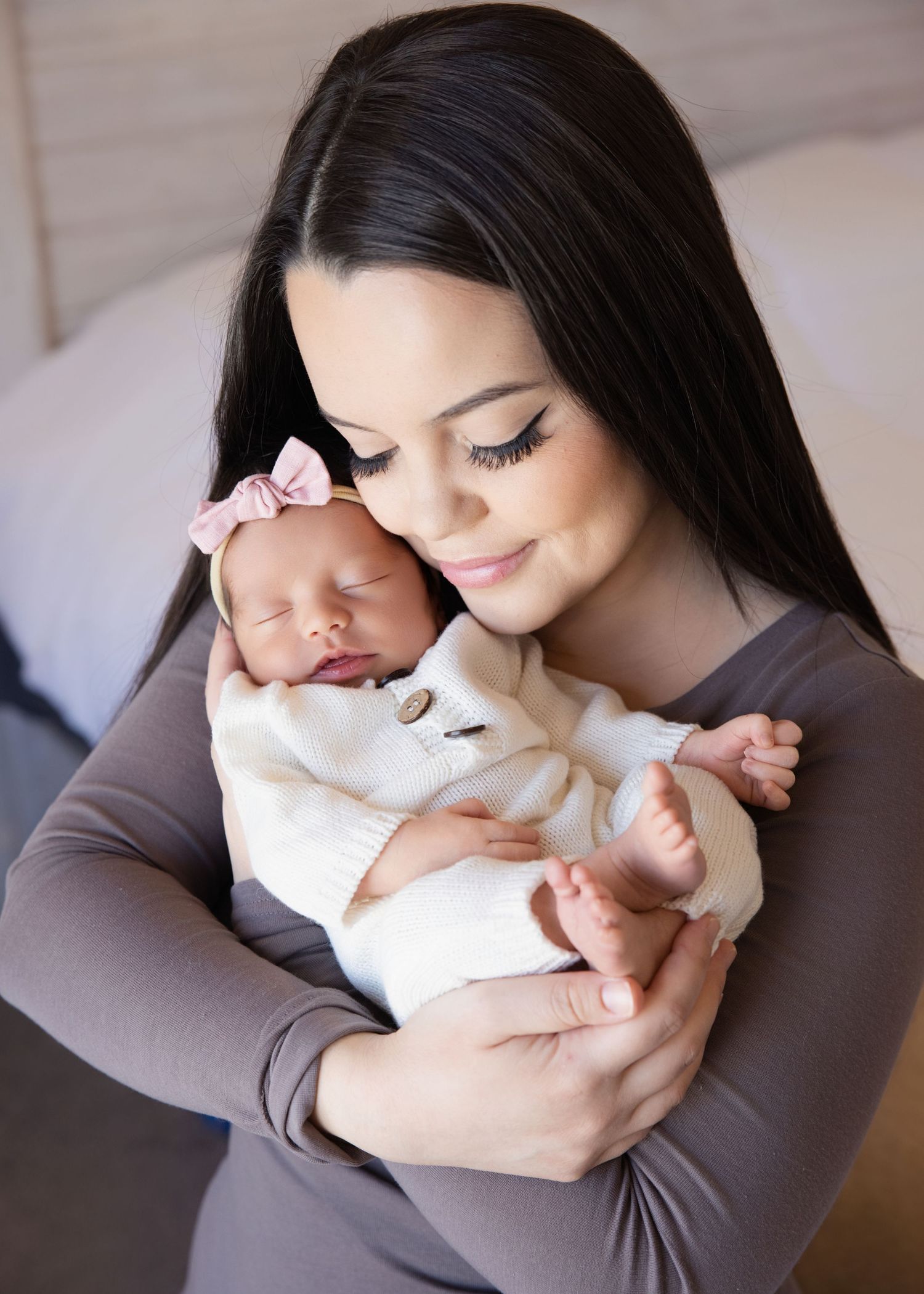 A young mother with dark hair tenderly holds her sleeping newborn baby dressed in white with a pink bow headband.