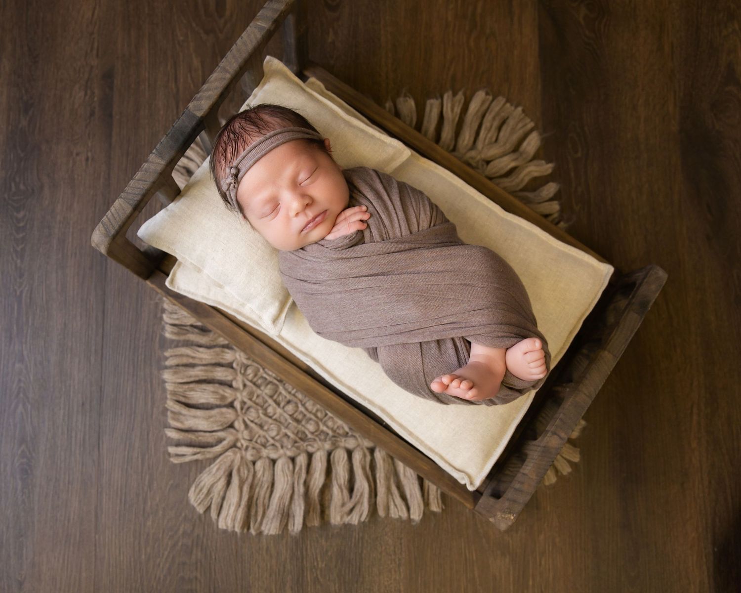 Newborn baby swaddled in brown wrap sleeping peacefully in a rustic wooden crate with cream pillow.