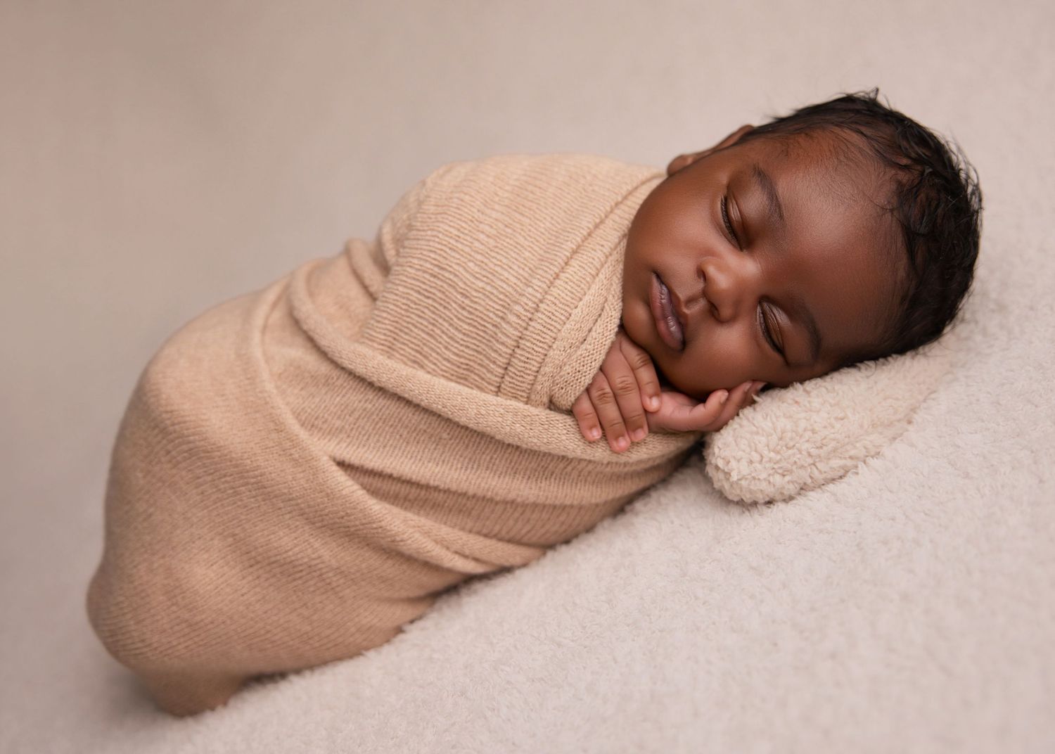 Sleeping newborn wrapped in soft beige fabric on a neutral blanket during a studio portrait.