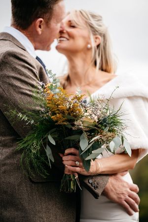Bride and groom share a romantic moment, bride holding a wildflower bouquet with greenery and yellow blooms on their wedding day.