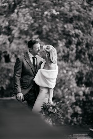 Black and white portrait of bride and groom gazing at each other outdoors with lush foliage background.