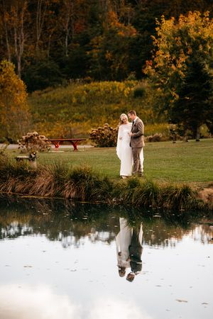 Bride and groom reflected in a still pond surrounded by vibrant autumn foliage during outdoor wedding portraits.
