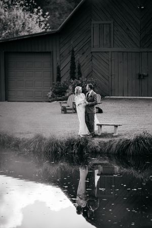 Black and white photo of a couple embracing by a pond near a rustic barn, their reflection visible in the still water.