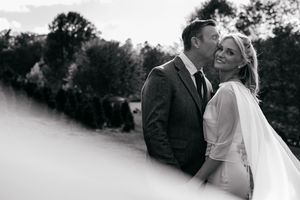 Black and white wedding photo of a smiling bride and groom embracing outdoors with lush greenery in the background.
