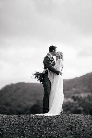 Black and white photo of a couple sharing a kiss on their wedding day with mountain scenery behind them.