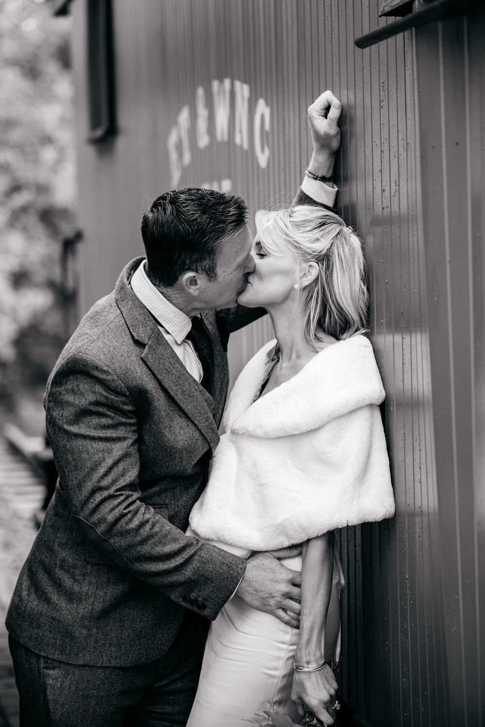 Black and white photo of bride and groom kissing against a vintage red train car on their wedding day.