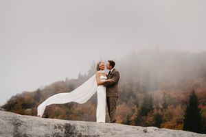 Bride and groom embrace on misty mountain rock, autumn foliage and fog-covered hills behind them.