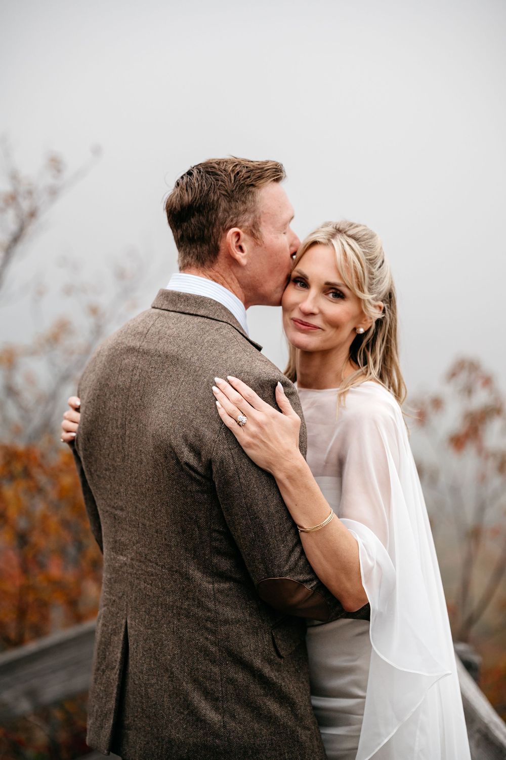 A groom in a brown tweed suit kisses his bride wearing a white cape dress outdoors in autumn.