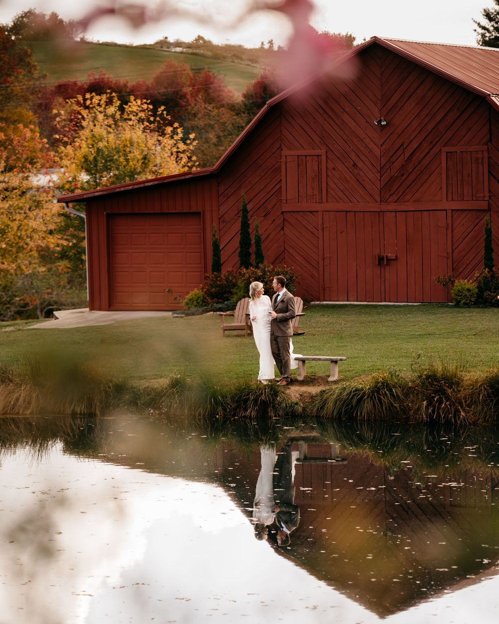 Couple standing by a pond reflecting a rustic red barn surrounded by vibrant fall foliage at a farm wedding.