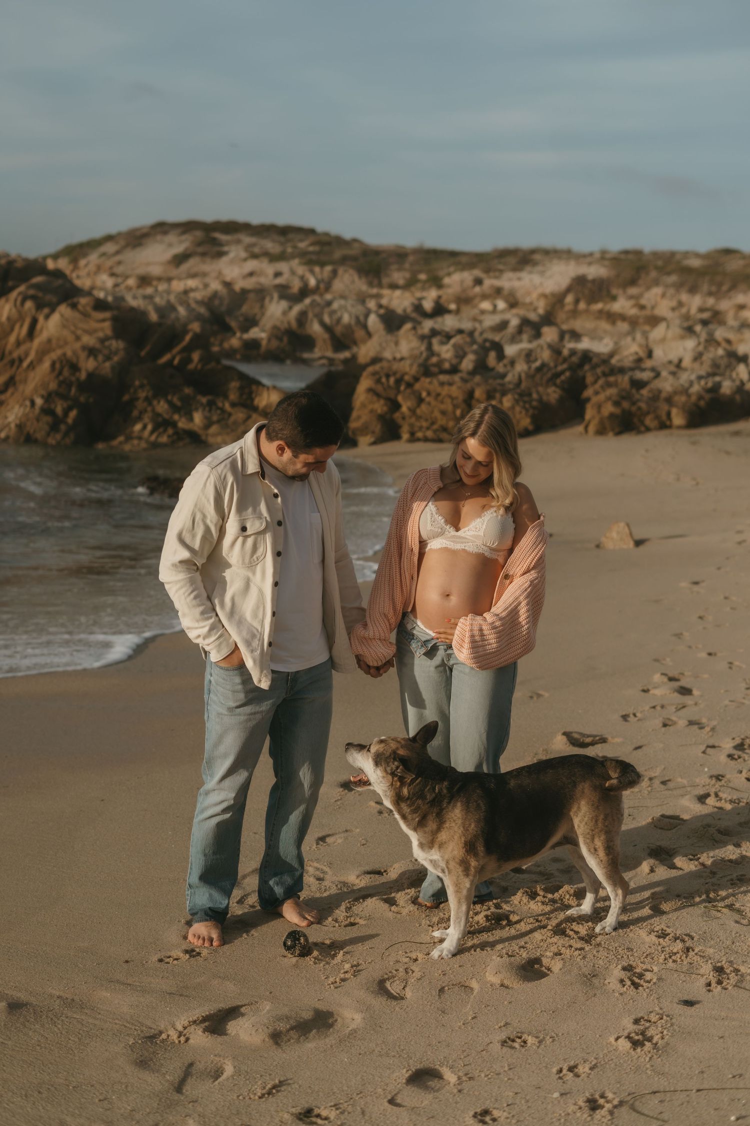 Pregnant couple holding hands on a sandy beach with their dog, rocky cliffs and ocean waves in the background.