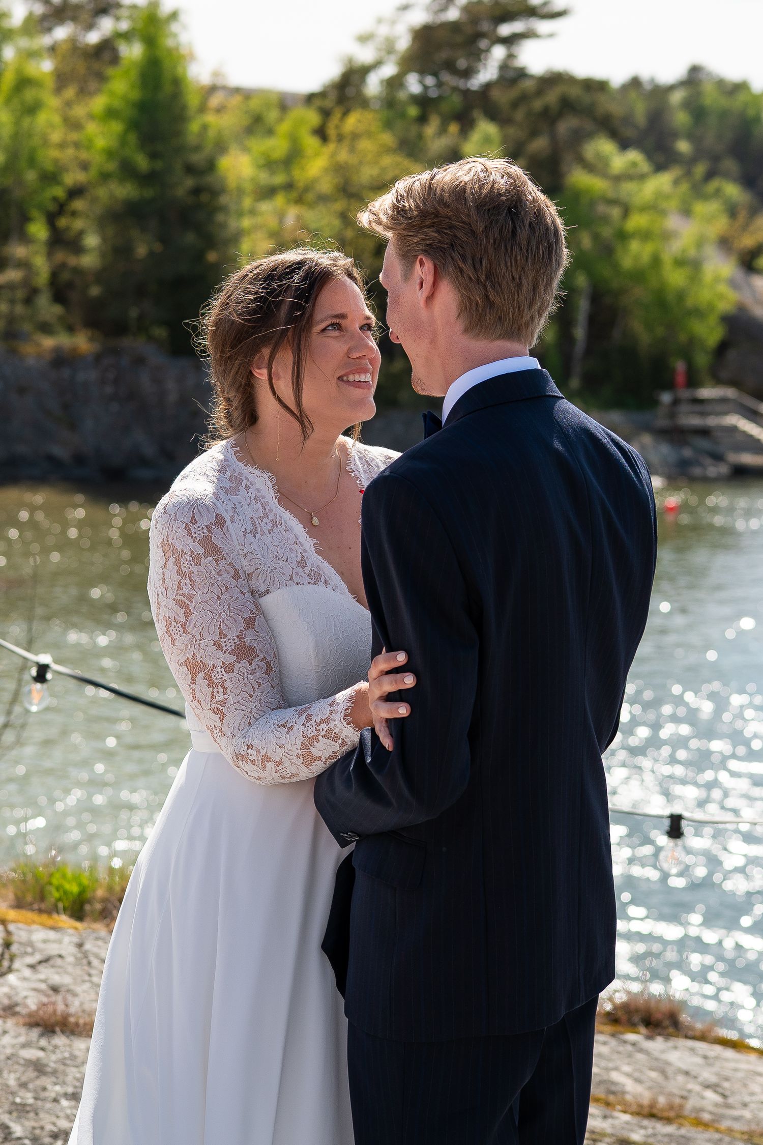 Bride in lace wedding dress and groom in navy suit embrace outdoors by a sunlit lake on their wedding day.