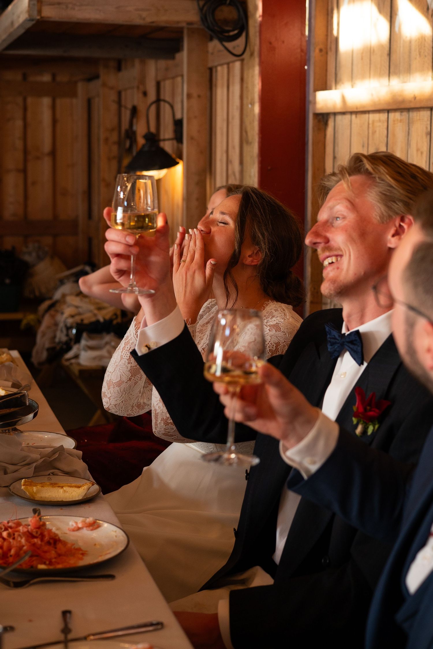 Bride and groom laughing and raising wine glasses at wedding reception dinner table in rustic venue.
