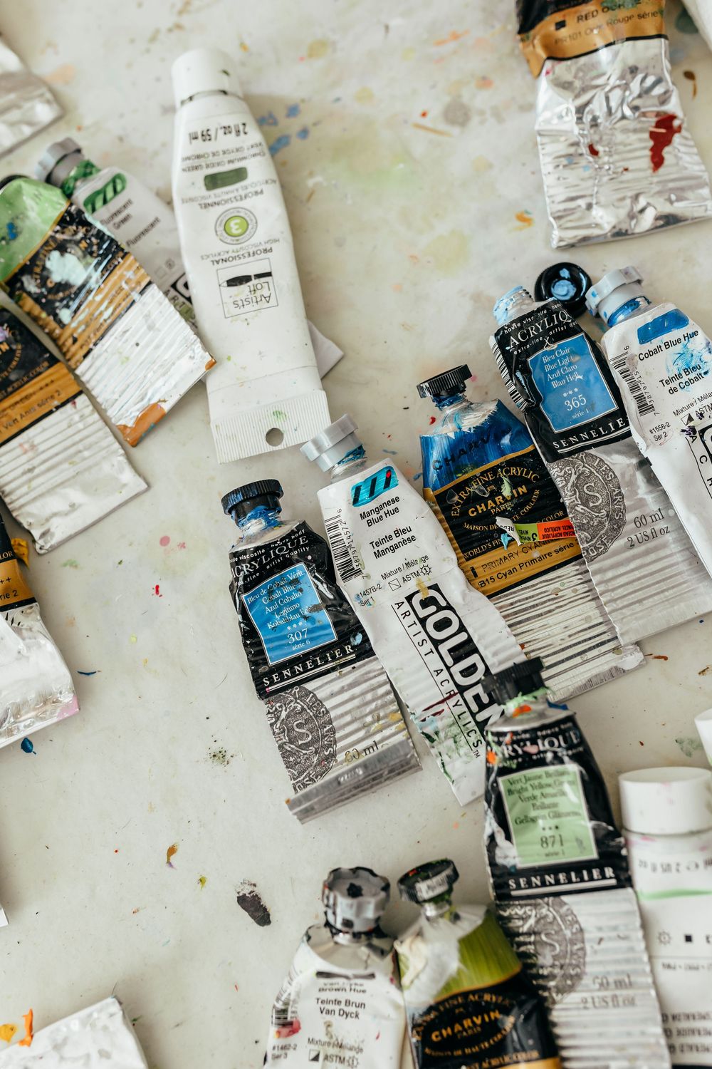 Various artist paint tubes including Golden, Sennelier, and Charvín brands scattered on a paint-stained white surface.