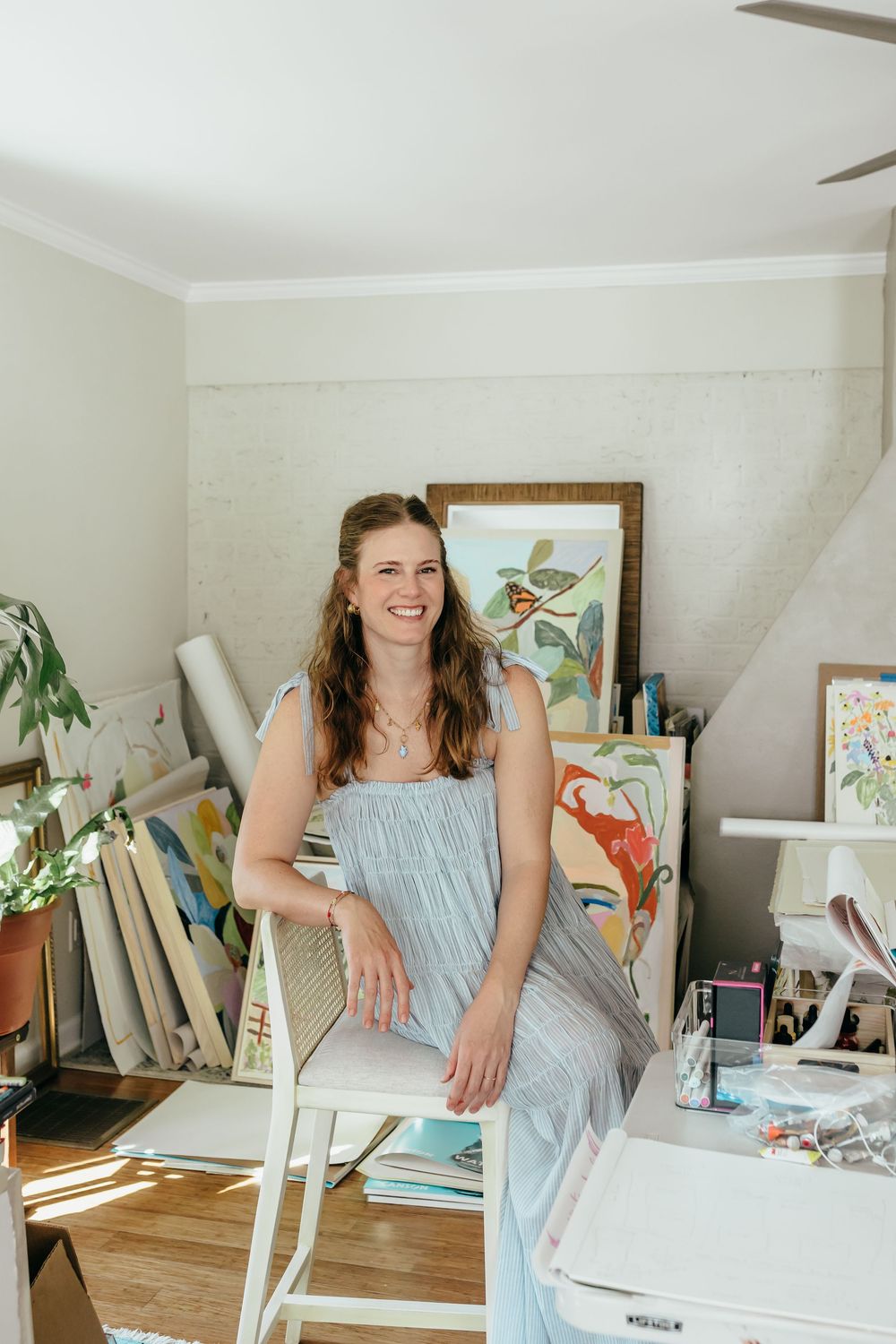 Smiling woman in light blue dress sits in art studio surrounded by colorful paintings and canvases.