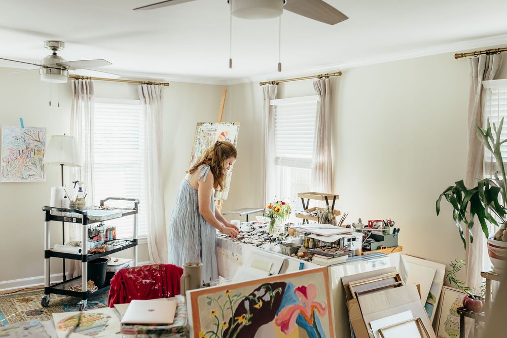 Artist woman working at a cluttered art studio table surrounded by colorful paintings and canvases in a bright room.