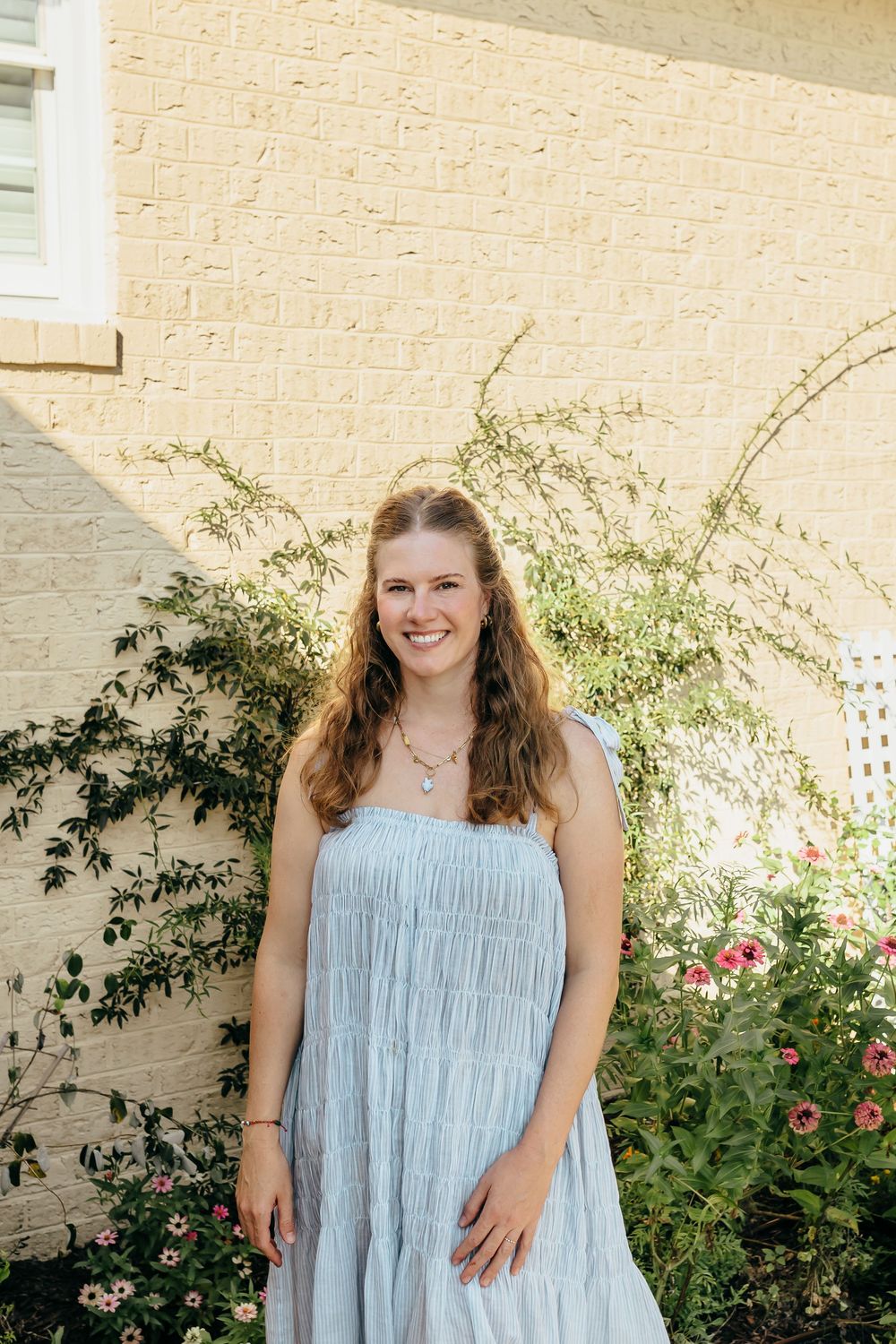 Young woman in light blue sundress smiling outdoors against brick wall with flowering garden plants.