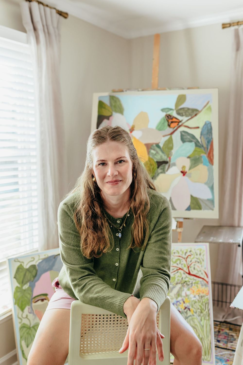 A smiling woman in a green cardigan sits in a cane chair in her art studio surrounded by colorful botanical paintings.