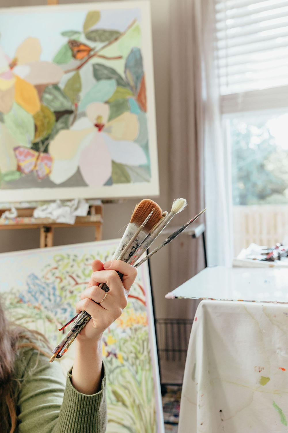 Artist holding multiple paint brushes in a bright studio with colorful floral paintings on easels in the background.