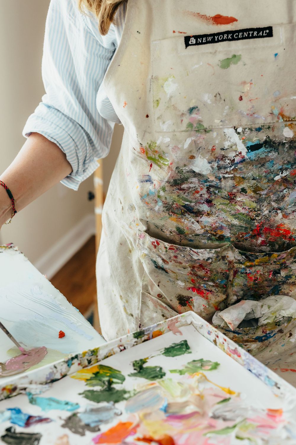 Artist in paint-splattered New York Central apron holds a colorful palette, showcasing vibrant mixed media painting supplies.