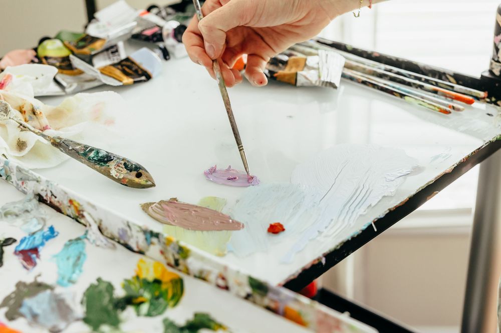 Artist mixing paint colors on a messy palette with a fine brush in a bright art studio.
