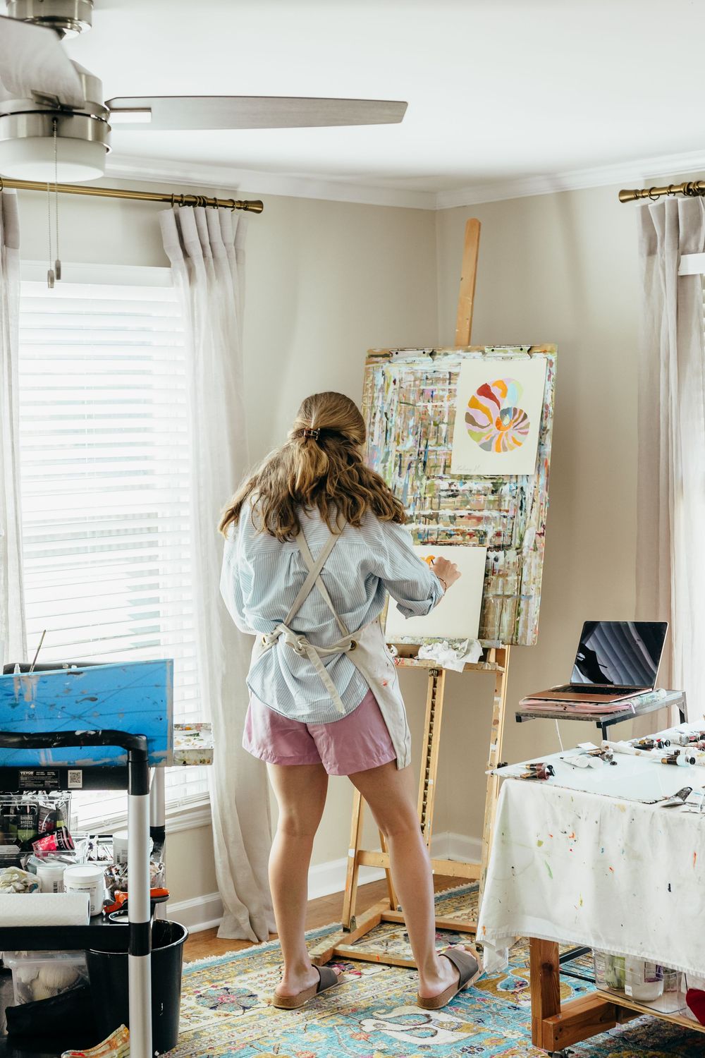 Young girl painting on canvas at wooden easel in bright home art studio with colorful palette and art supplies.