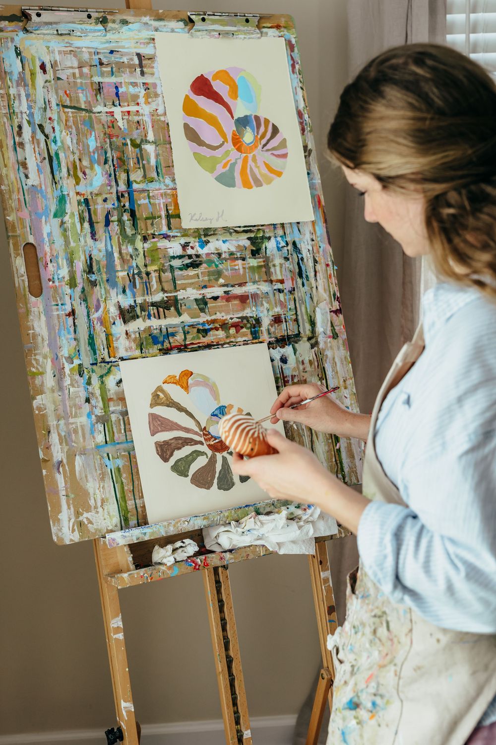 Artist painting colorful nautilus shell designs on canvas displayed on a paint-splattered wooden easel in a bright studio.