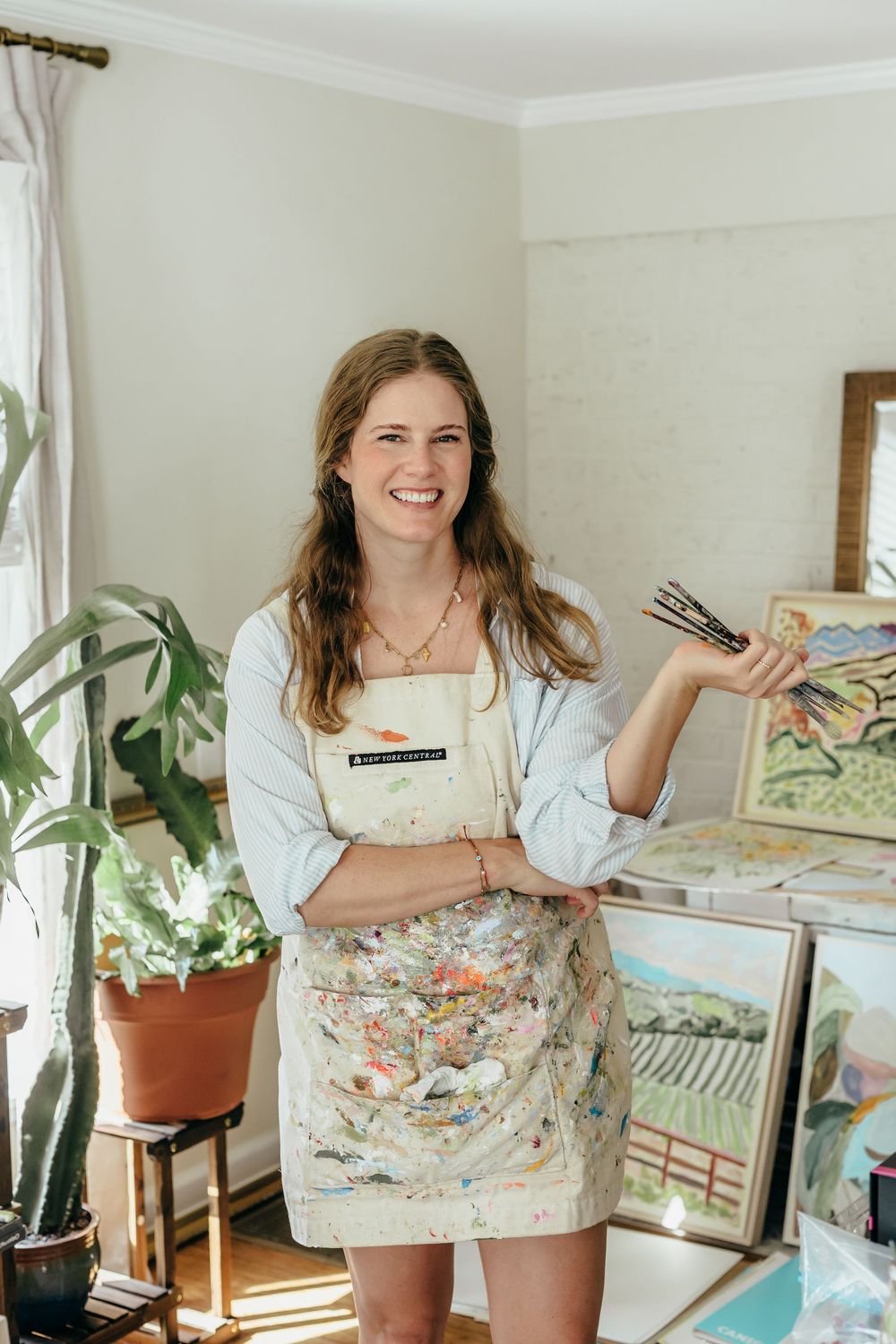Smiling female artist in a paint-splattered apron holds brushes in a bright studio surrounded by colorful paintings and plants.