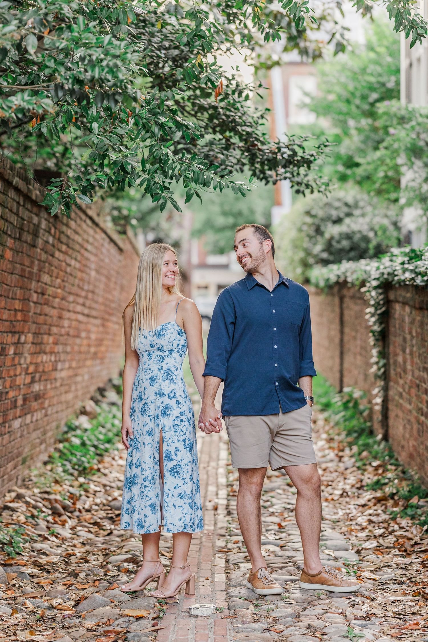 Couple holding hands and smiling on a brick path lined with ivy walls and lush greenery.
