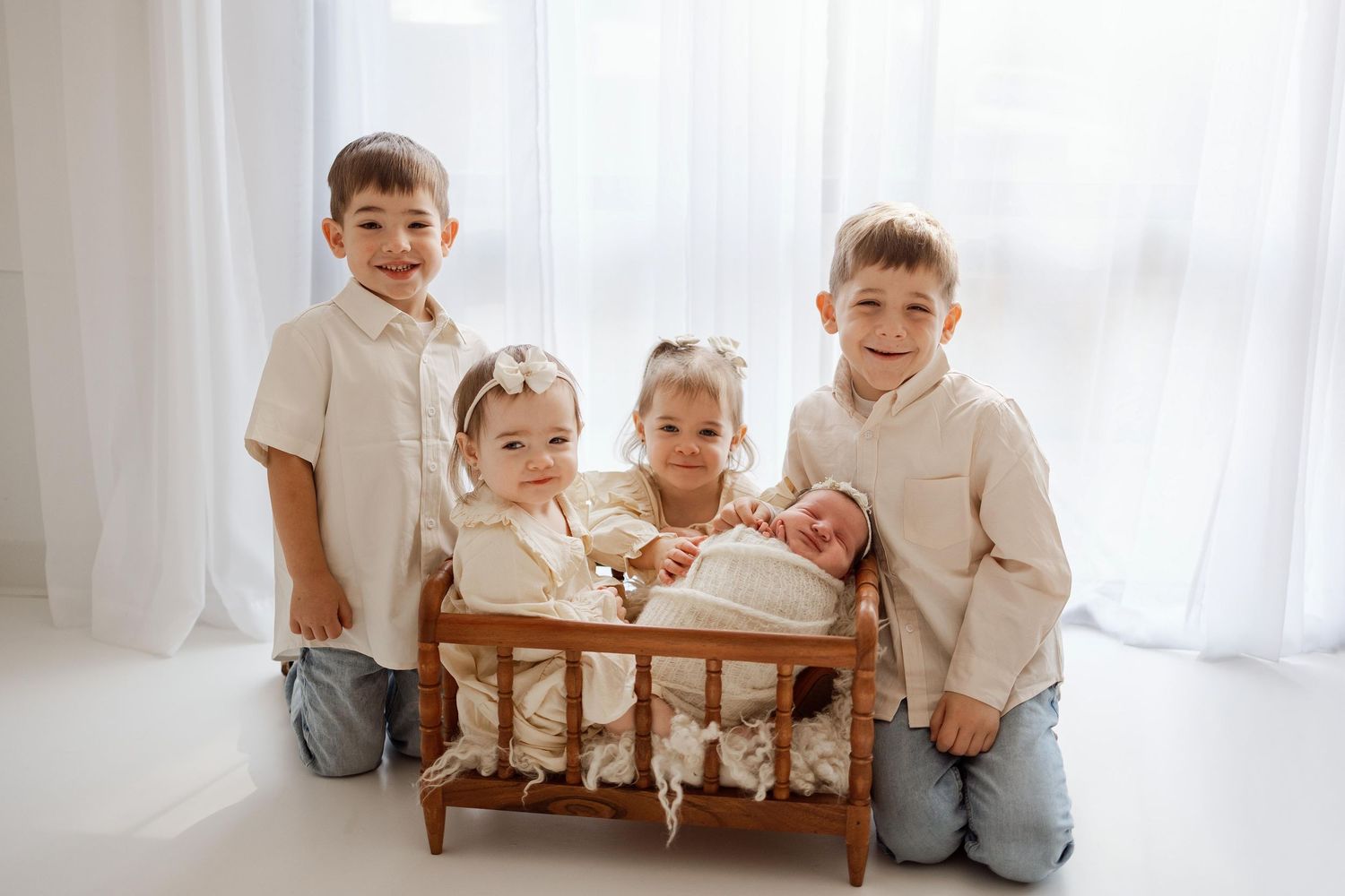 Four young siblings pose with a swaddled newborn in a wooden doll bed during a professional family photo session.