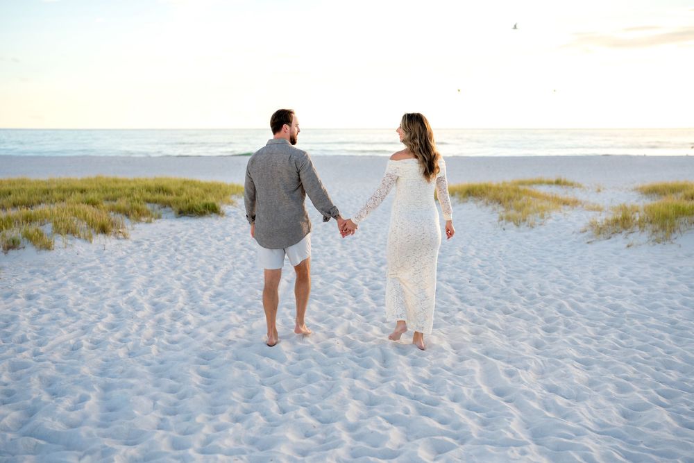 Couple holding hands walking on white sand beach at sunset, woman in white dress and man in gray shirt.