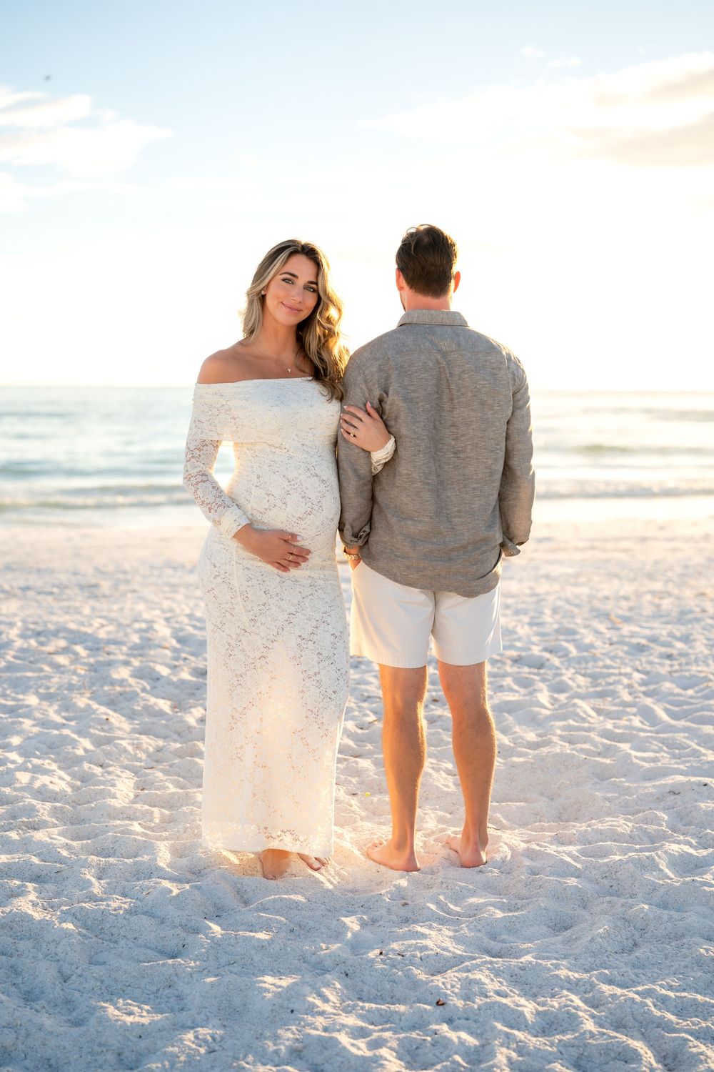 Pregnant woman in white lace dress poses with partner on sandy beach at sunset.