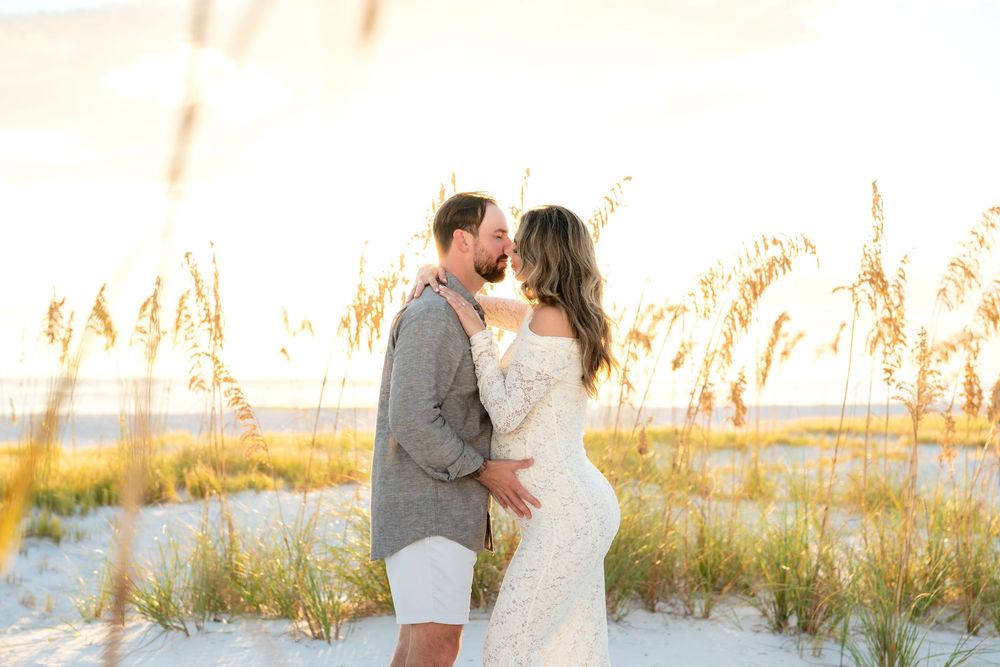 Couple sharing a romantic kiss on a sandy beach at golden hour, surrounded by sea oats, bride in lace gown.