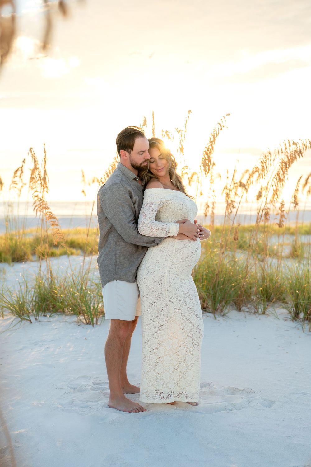 Couple embracing on white sand beach at golden hour, pregnant woman in lace gown with sea oats backdrop.
