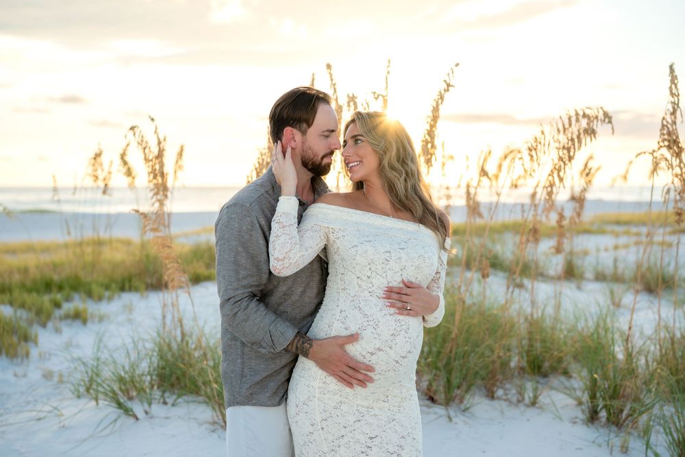 Couple sharing a tender moment on a white sand beach at sunset, woman in white lace maternity gown cradling her bump.