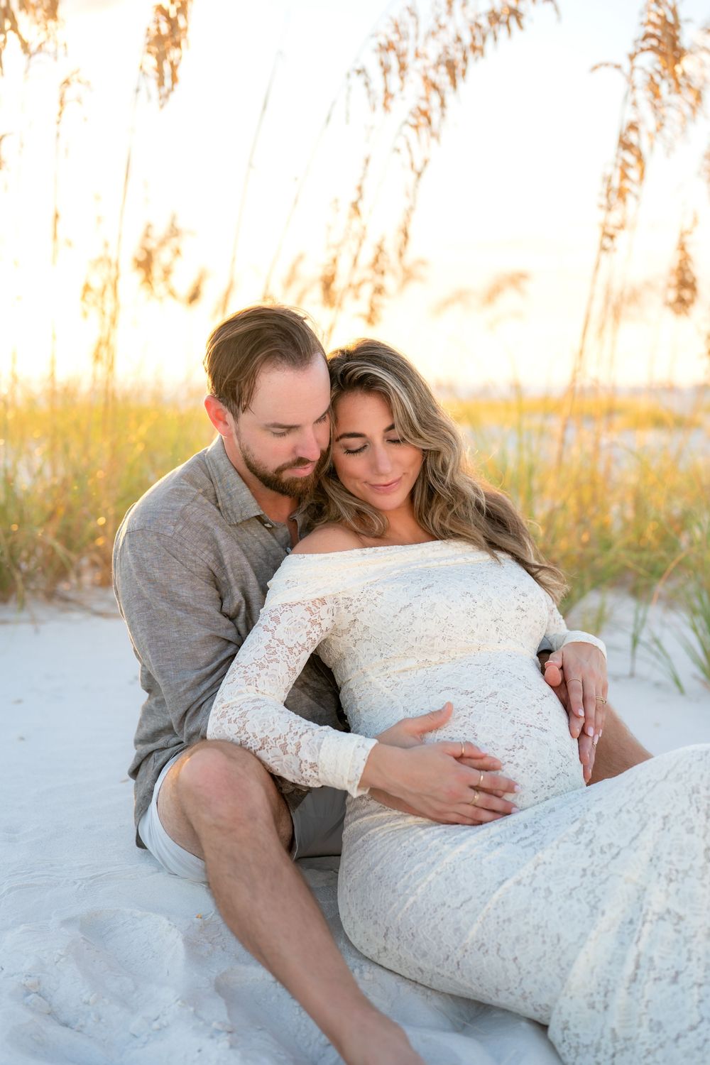 Couple sitting on beach sand dunes at sunset, woman in white lace maternity dress, man embracing her from behind.