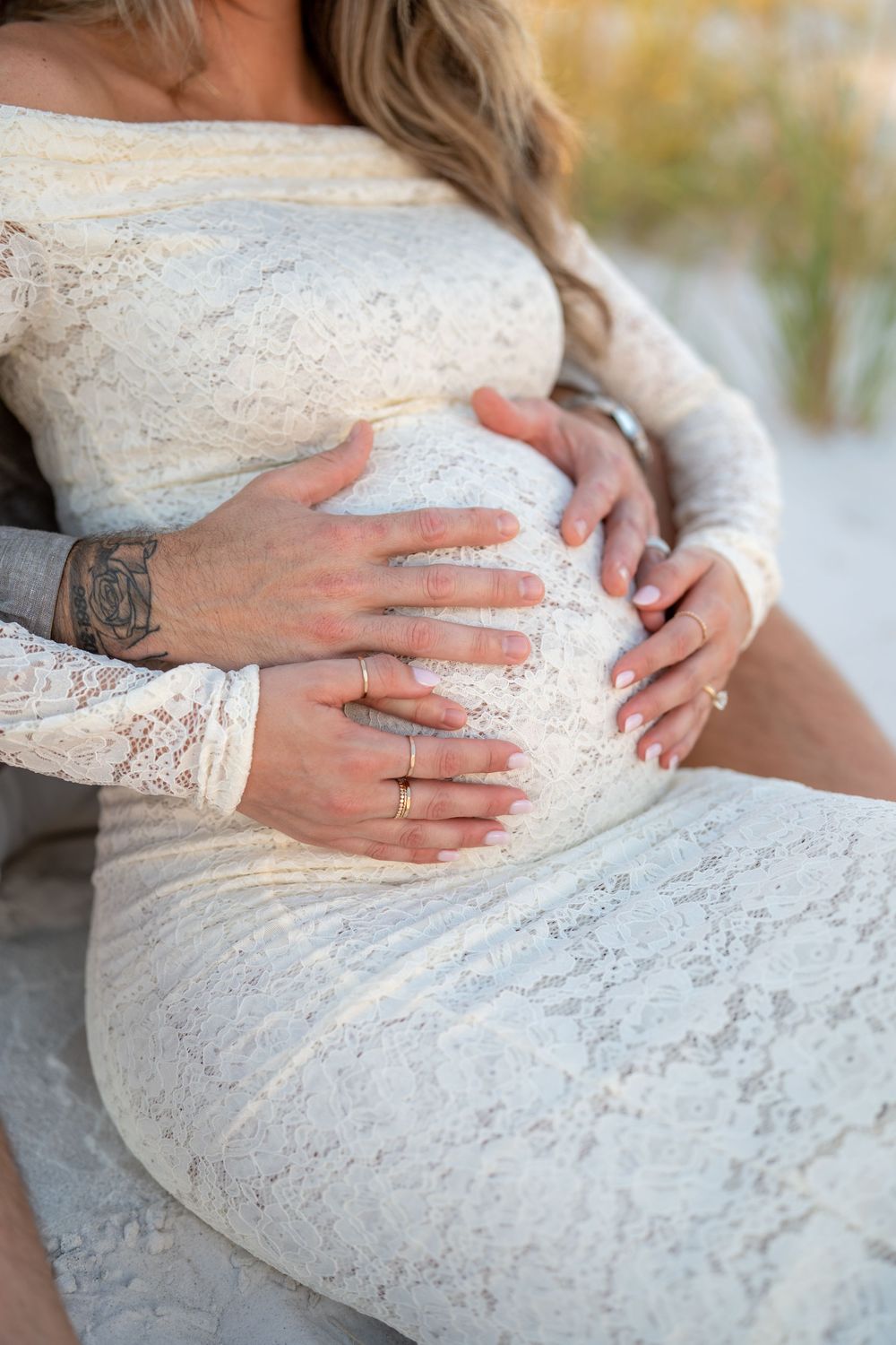 Pregnant woman in white lace dress with partner's tattooed hands cradling baby bump during maternity photo session.