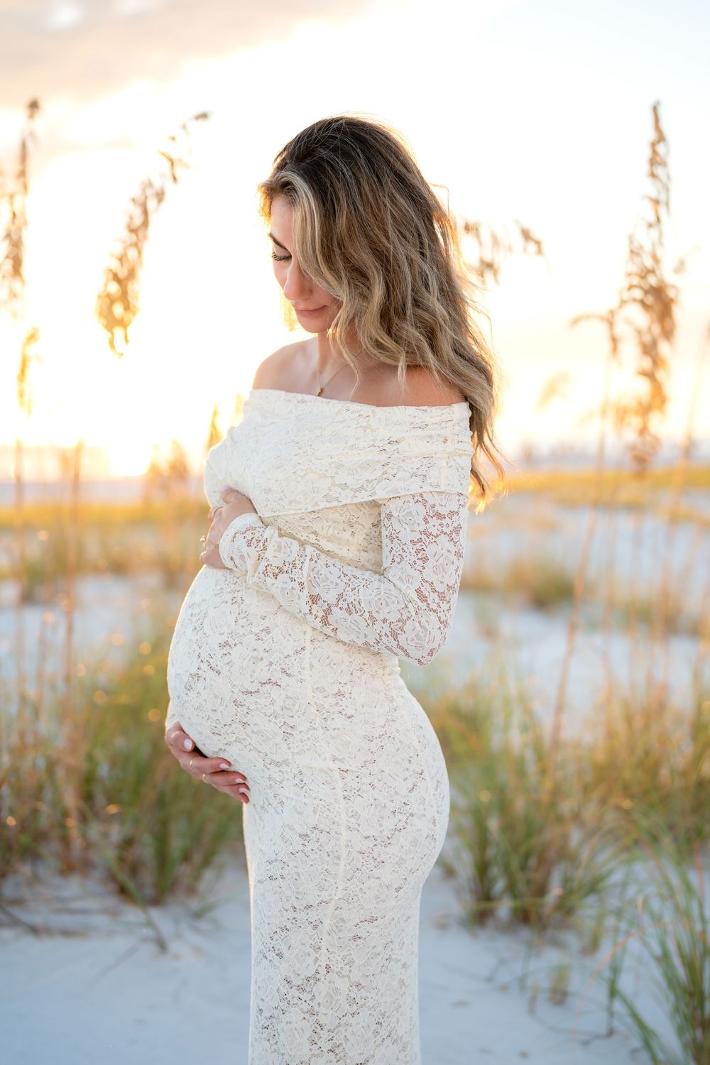 Pregnant woman in white lace off-shoulder maternity gown cradling baby bump at beach sunset.