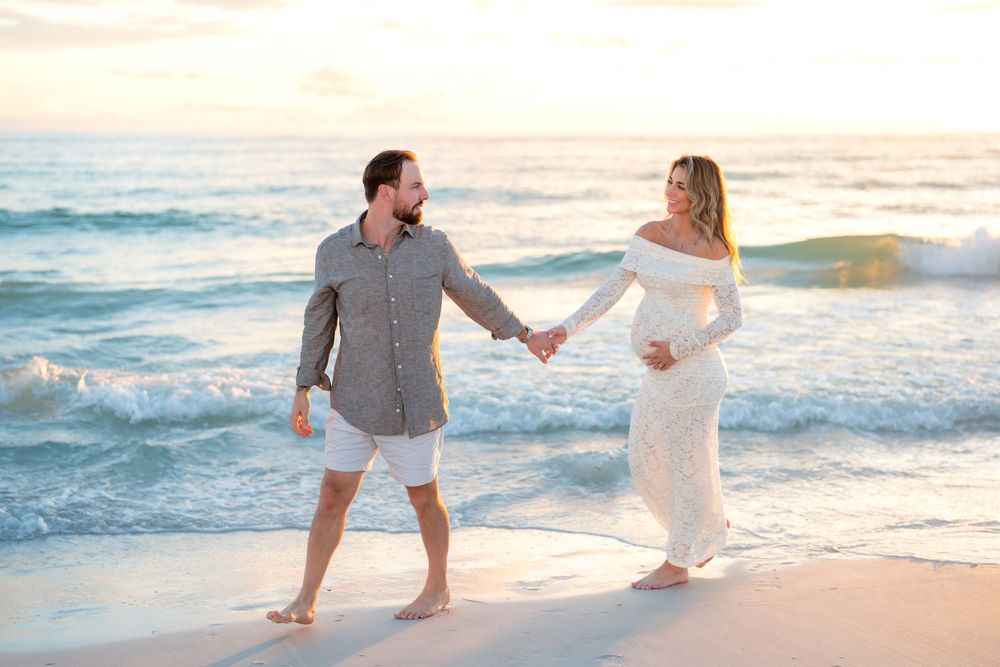 Couple holding hands walking on a sandy beach at sunset, woman in white maternity gown, man in gray shirt.
