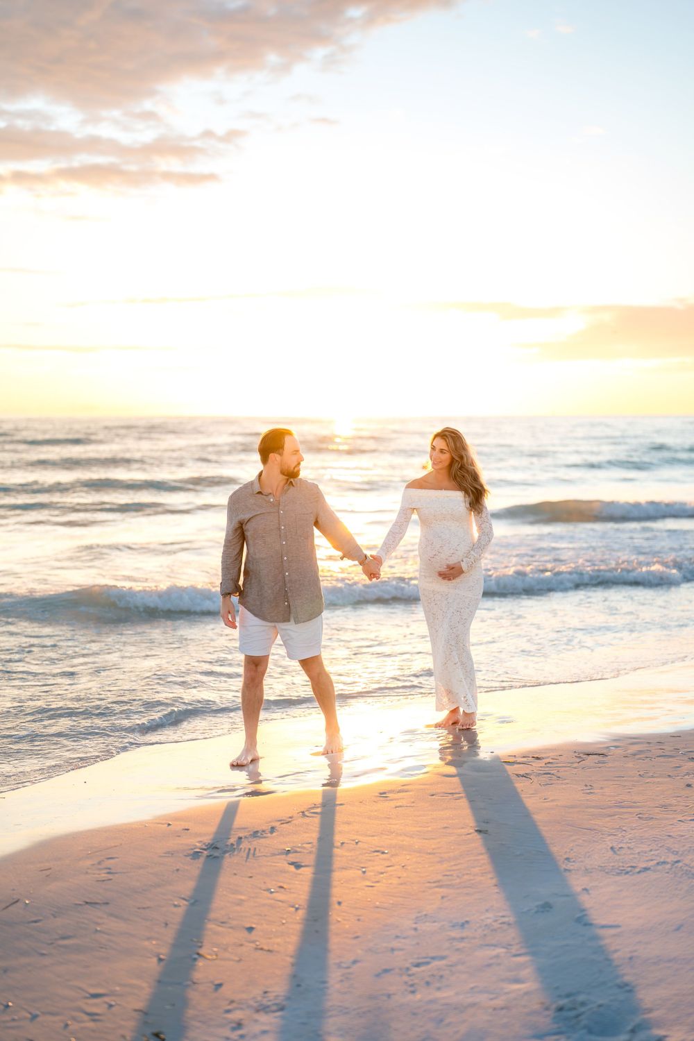 Pregnant couple holding hands on a sandy beach at golden sunset, woman in white lace dress touching baby bump.