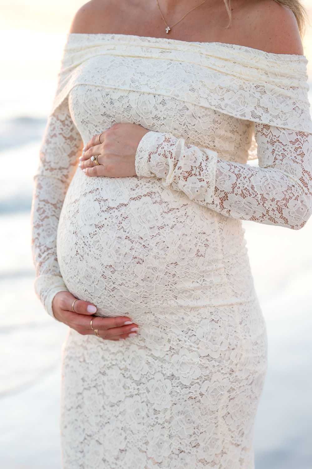 Pregnant woman in elegant off-shoulder white lace maternity gown cradling baby bump at beach.