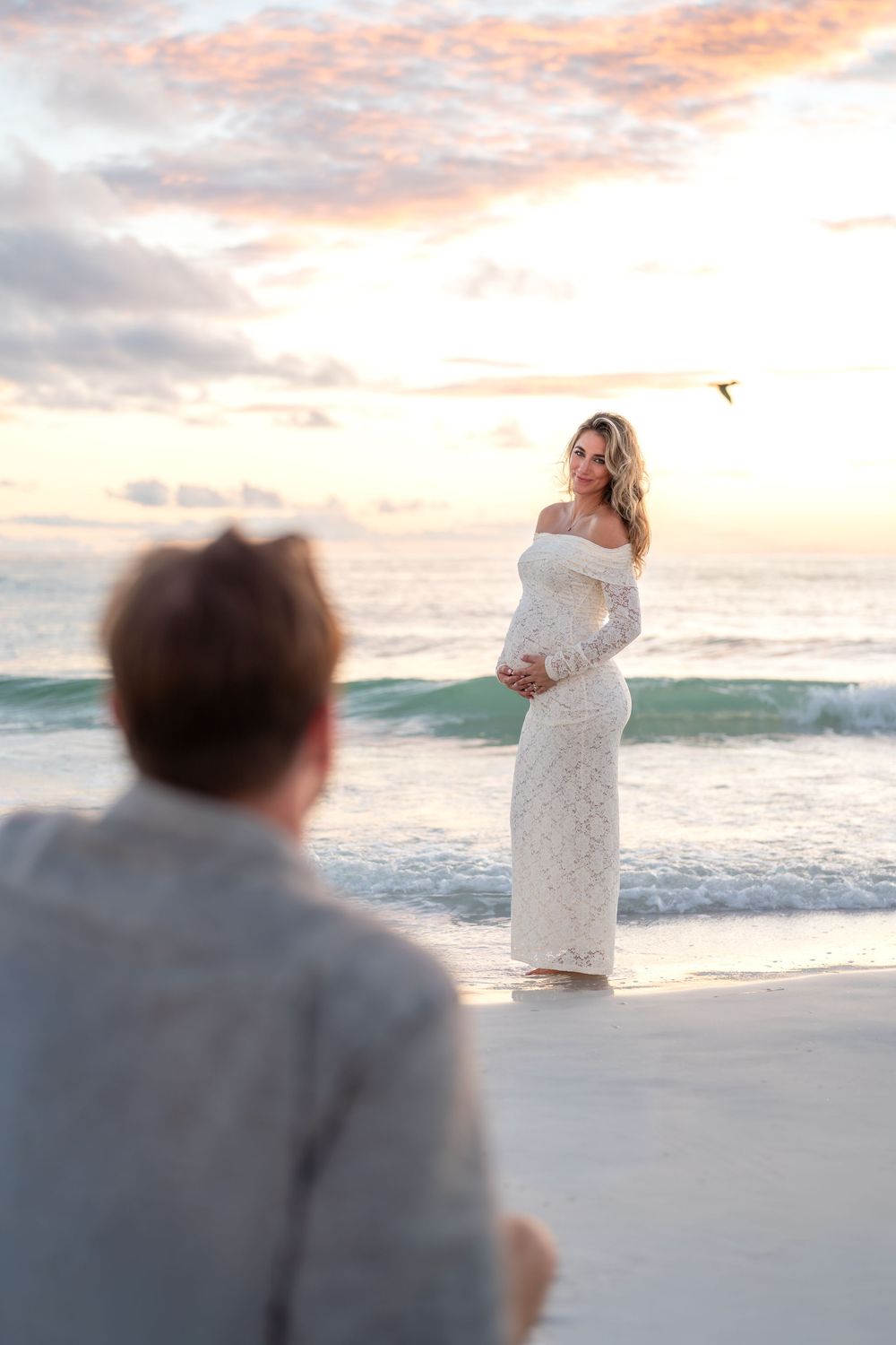 Pregnant woman in white lace gown poses on beach at sunset while partner watches, ocean waves in background.