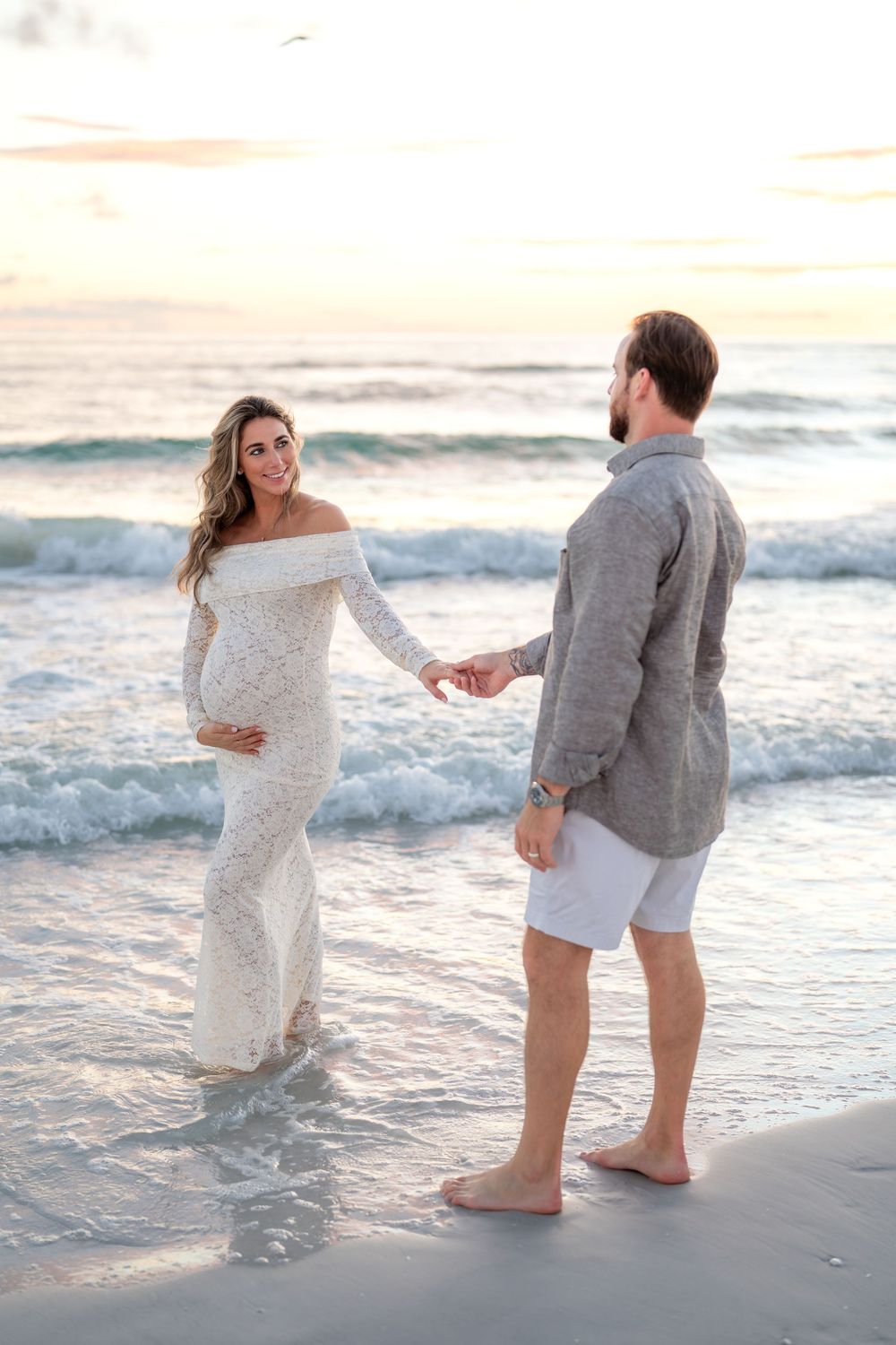 Pregnant woman in white lace gown holds hands with man on beach at sunset.
